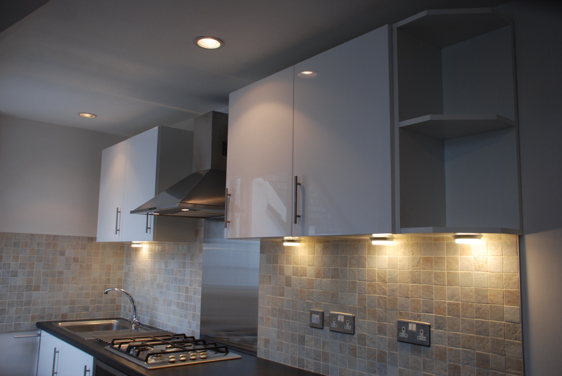 Kitchen detail showing stainless-steel extractor hood, gloss-white wall cabinets, stone-effect mosaic tile backsplash, and under-cabinet LED lighting over the gas hob.