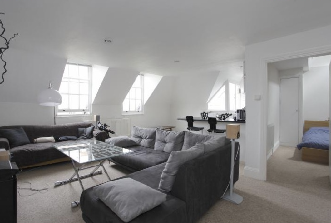 Open-plan reception room with two grey upholstered sofas, glass coffee table, dormer windows letting in natural light, kitchen bar with stools on the right, and the bedroom doorway at the rear.